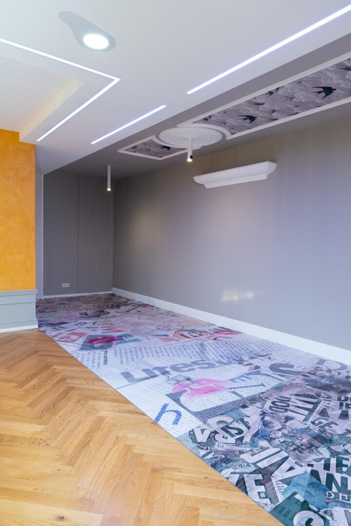 Moderner, minimalistischer Raum mit grauen Wänden, geometrischer Deckengestaltung, orangefarbener Akzentwand und einem Boden mit einer Collage aus Zeitungsdrucken und Grafiken. Holzparkett geht in den gemusterten Bereich über.