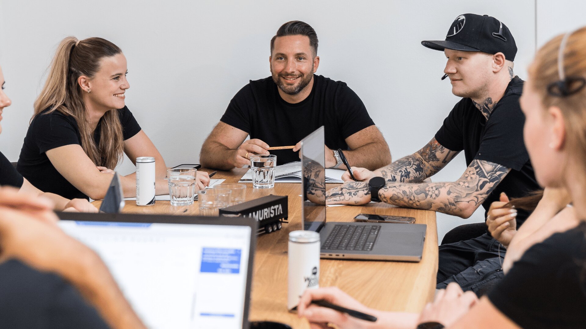 Sechs Personen sitzen um einen Holztisch und halten eine Besprechung ab. Laptops, Getränke und Notizblöcke stehen bereit. Die Gruppe wirkt engagiert und lächelnd, eine Person trägt einen Hut und ihre Tattoos sind deutlich zu sehen.