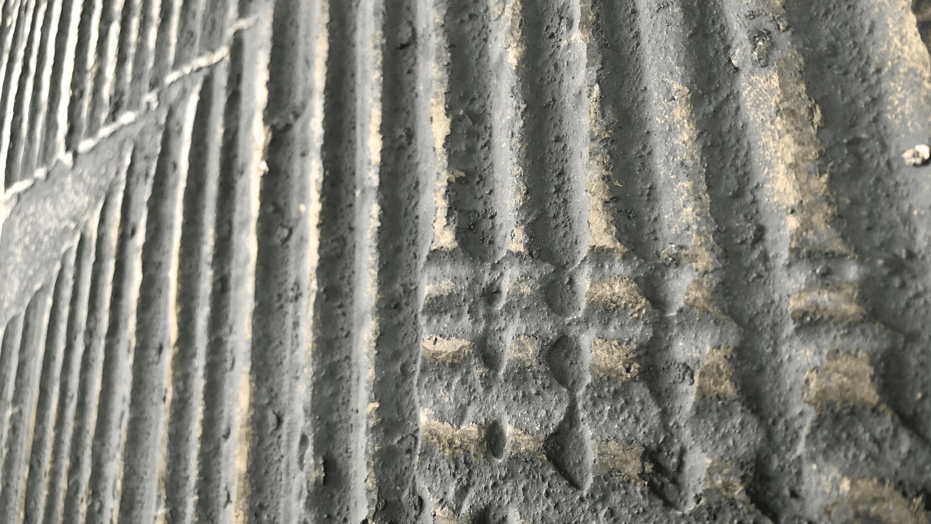 Nahaufnahme einer strukturierten, gerippten Betonwand mit vertikalen Rillen und rauer, unebener Oberfläche. Die Wand wirkt abgenutzt, mit kleinen Verfärbungen und angesammeltem Staub oder Schmutz.