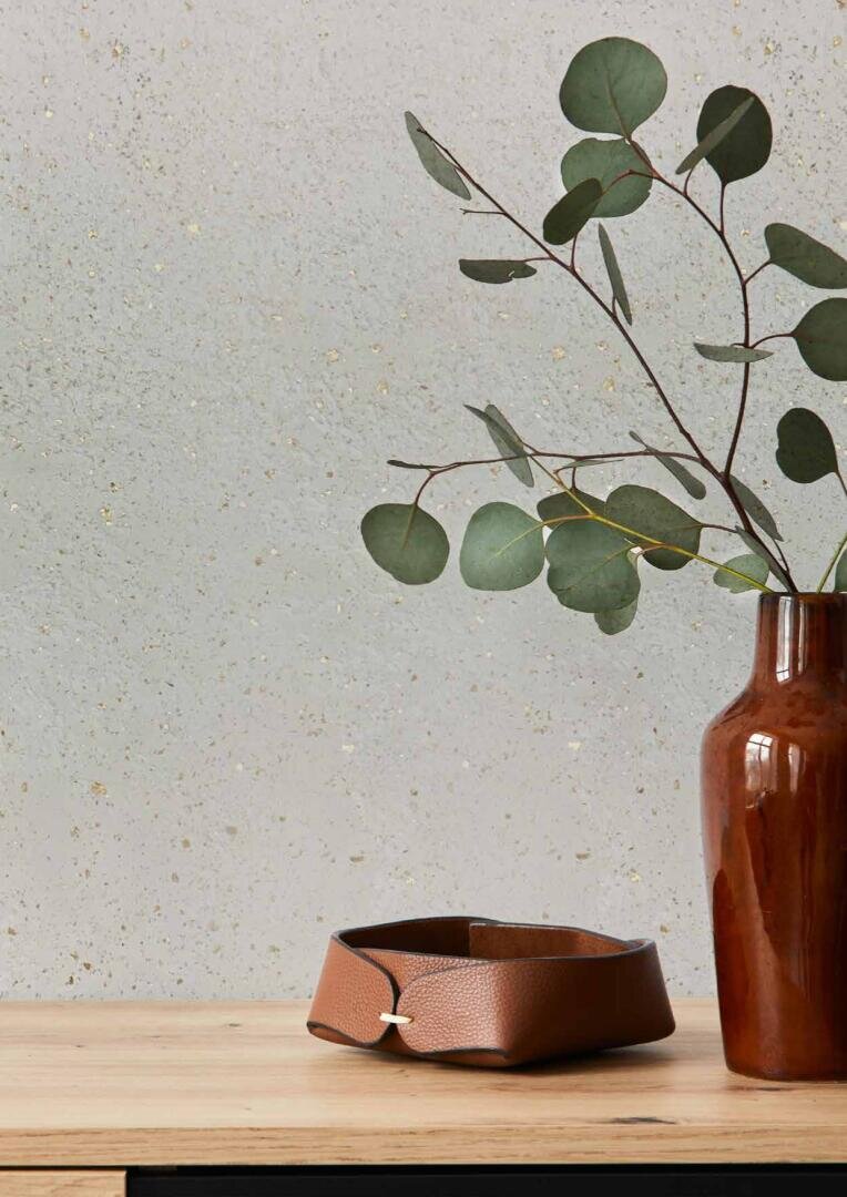 A brown vase with green eucalyptus branches sits on a wooden surface next to a folded brown leather accessory against a light gray textured wall.