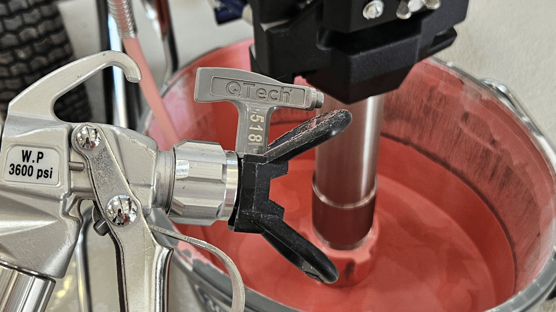Nahaufnahme einer Farbspritzpistole mit der Aufschrift „W.P. 3600 psi“ und „QTech“ neben einem mit rosa Flüssigkeit gefüllten Mischbehälter, im Hintergrund mischt ein Metallstab den Inhalt.