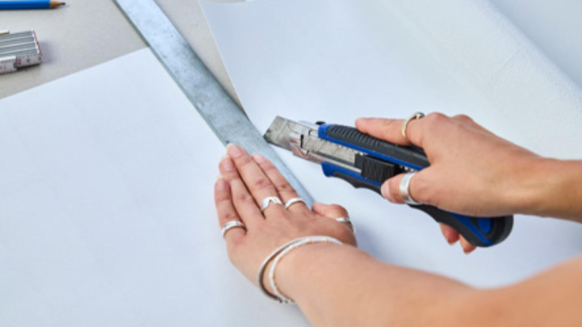 Hände schneiden mit einem Universalmesser und einem Metalllineal ein weißes Blatt Papier auf einer flachen Oberfläche aus. Im Hintergrund sind einige Schreibwaren zu sehen.