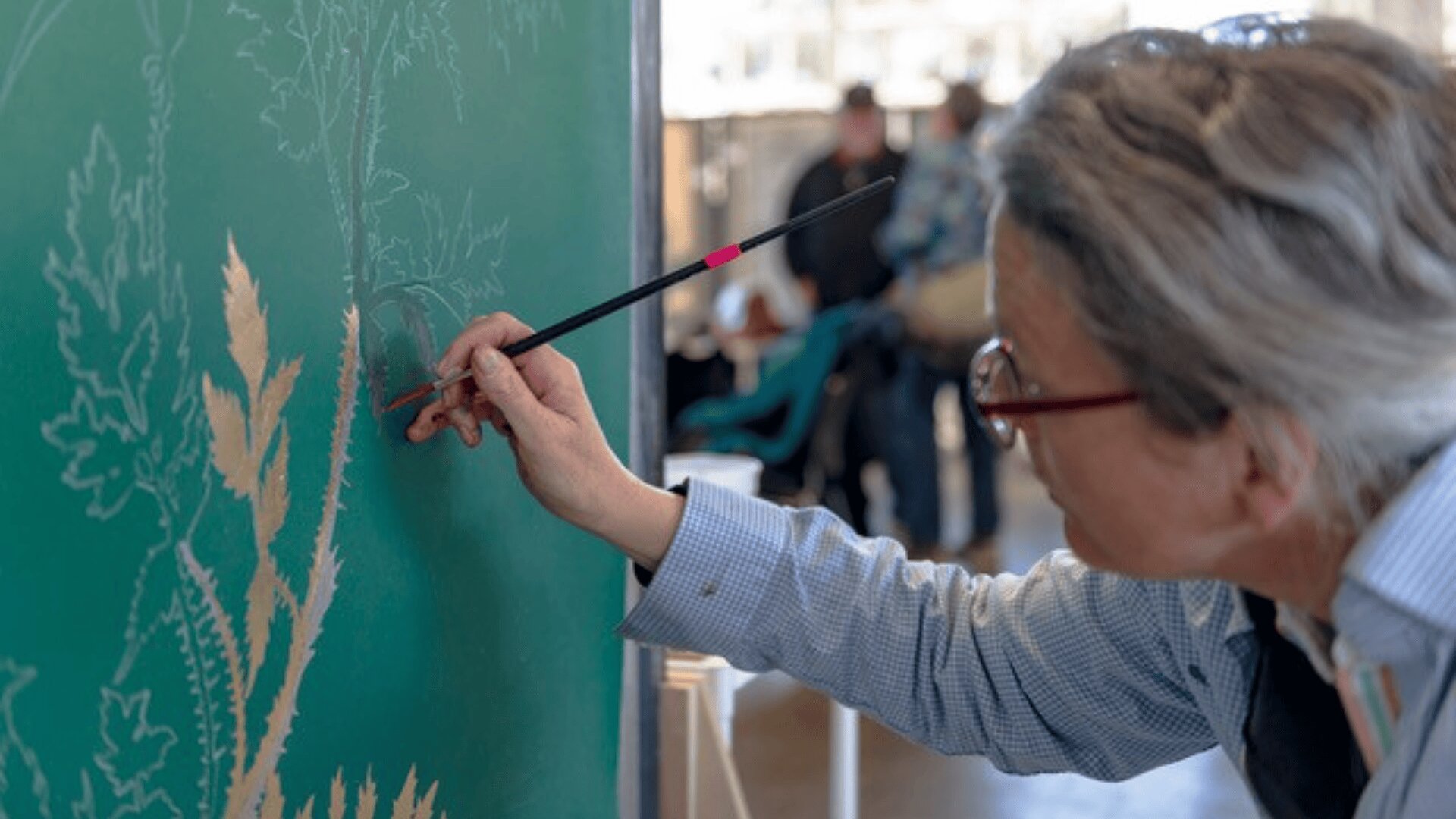 Eine Person mit grauem Haar und Brille malt detailreiche Blätter auf eine grüne Fläche und hält dabei einen feinen Pinsel in der Hand. Der Hintergrund ist leicht verschwommen und zeigt andere Personen und Aktivitäten.