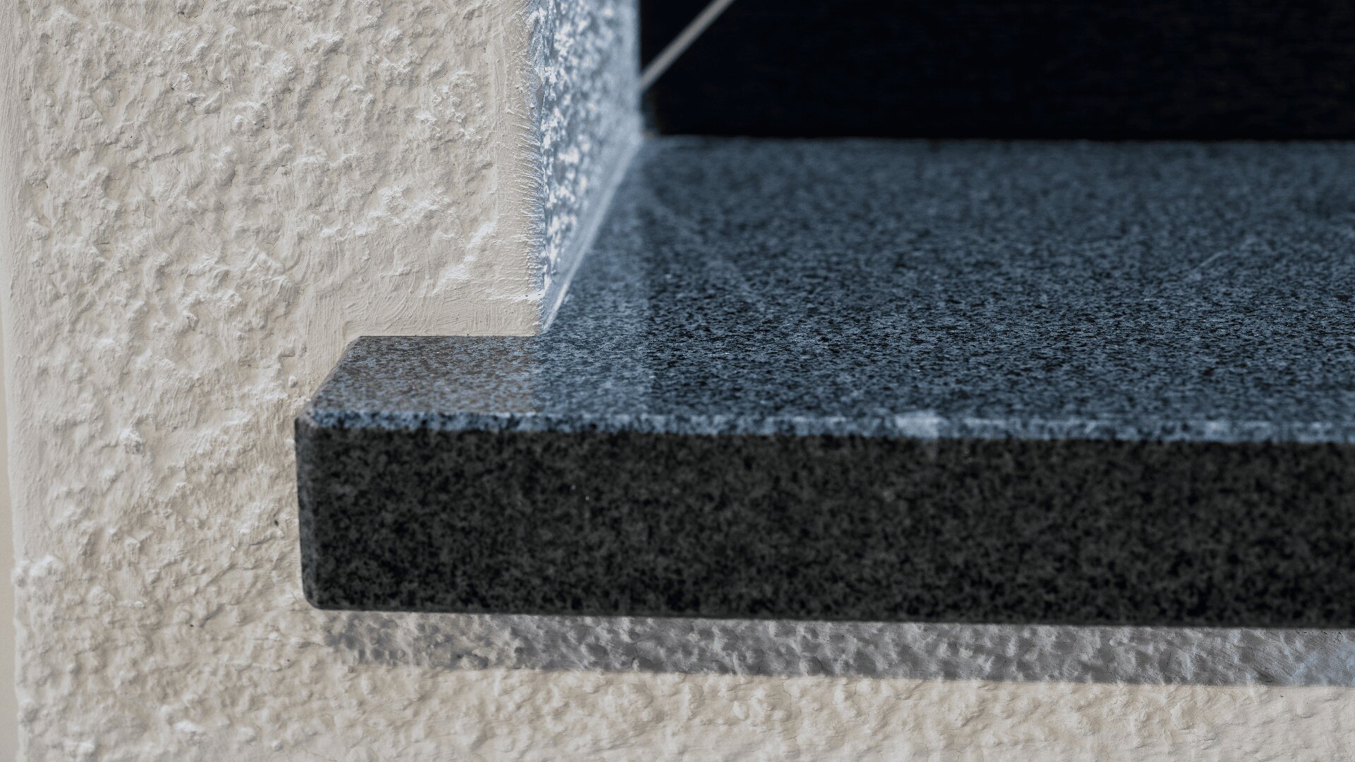 Nahaufnahme einer Fensterbank oder eines Fenstersimses aus dunklem Granit mit glatter, polierter Oberfläche und abgeschrägter Kante vor einer strukturierten, hellen Wand.