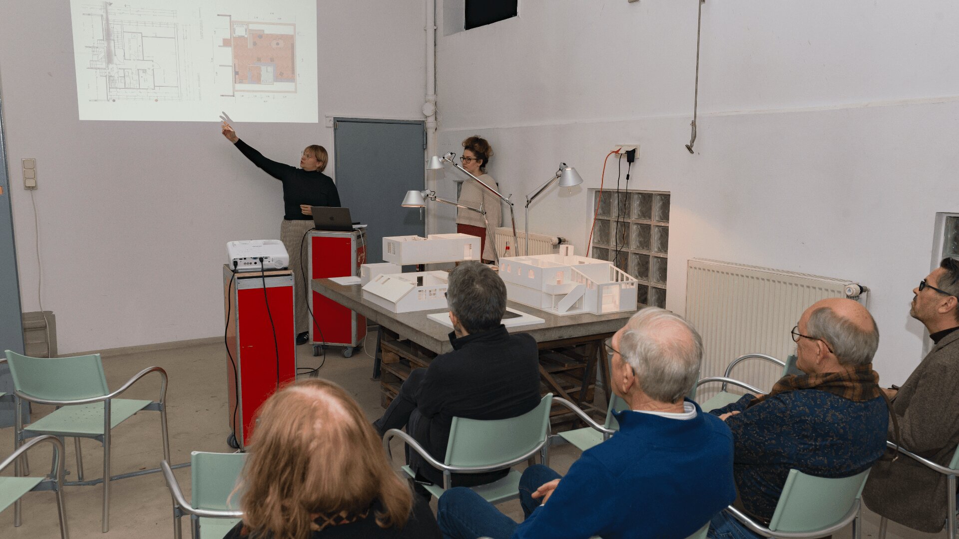 Zwei Personen präsentieren einem sitzenden Publikum in einem Raum Architekturpläne mit Folien und maßstabsgetreuen Modellen. Ein Vortragender zeigt auf eine Projektion an der Wand, während der andere neben einem Laptop steht.