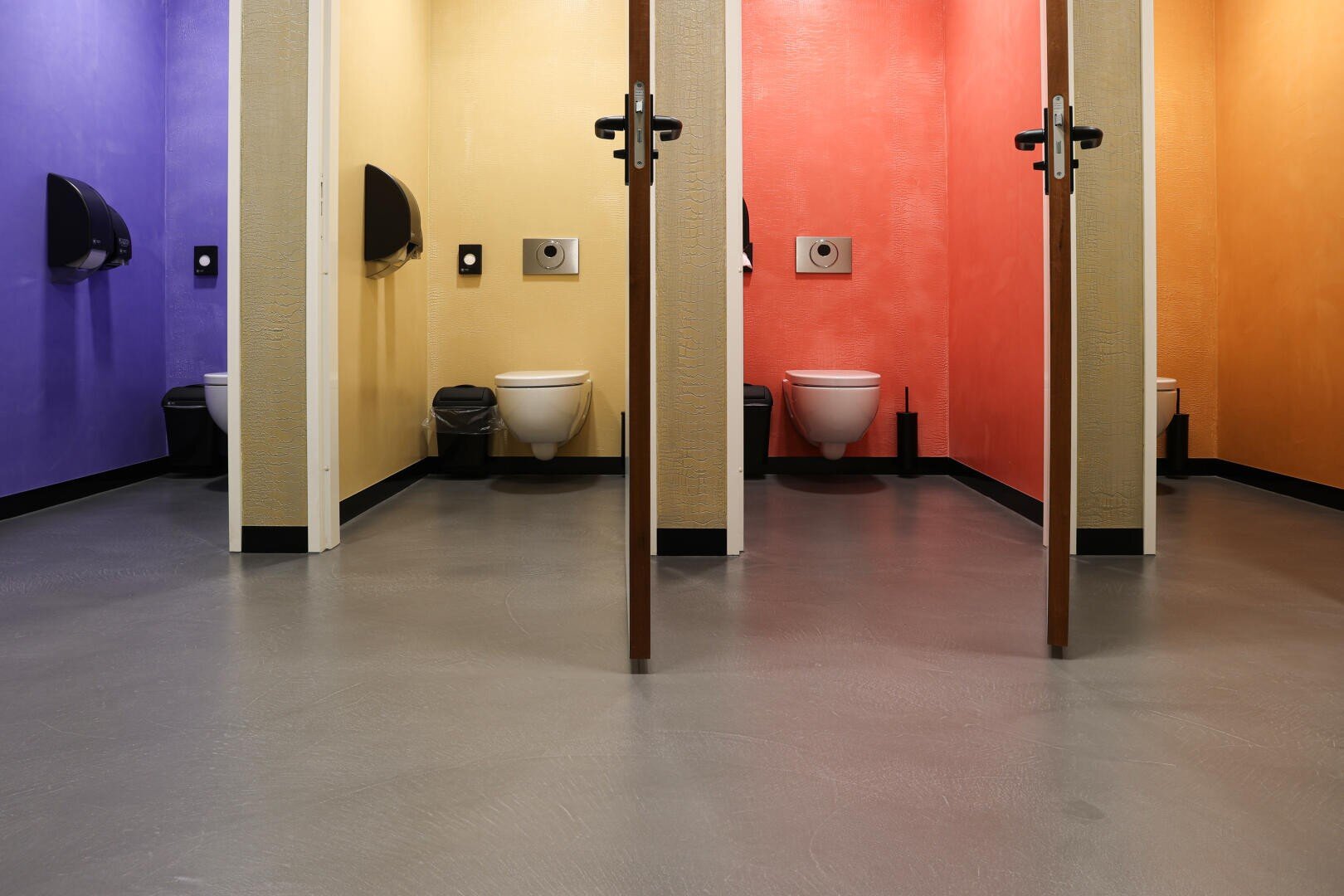 Drei Toilettenkabinen mit bunten Wänden: links lila, in der Mitte rot und rechts gelb. Jede Kabine verfügt über eine Toilette, einen Toilettenpapierspender und einen Mülleimer. Der Boden ist grau.