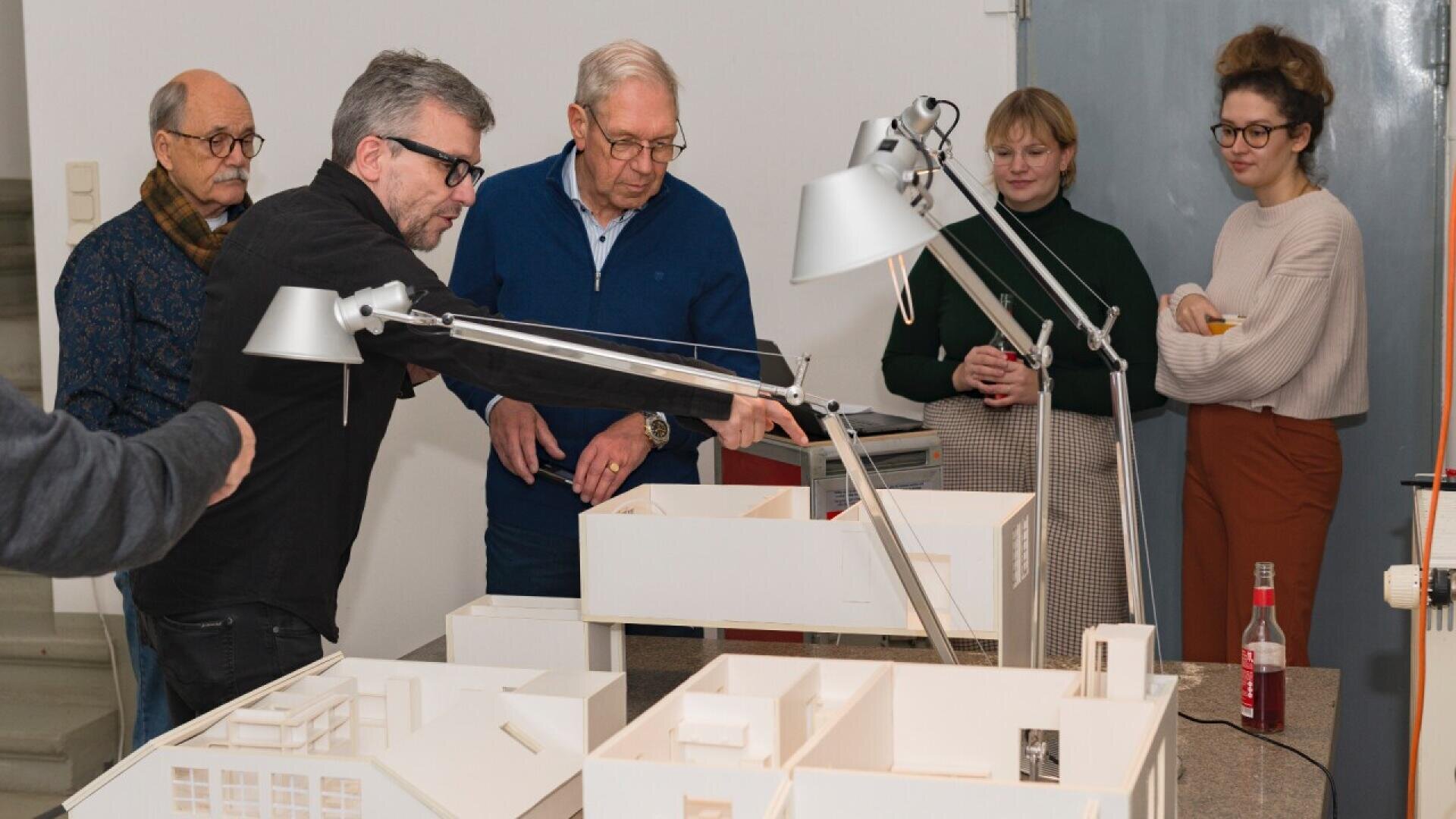 Five people stand around a table, examining and discussing detailed architectural models of buildings. Two men in front gesture towards the models, while three others observe attentively in the background.