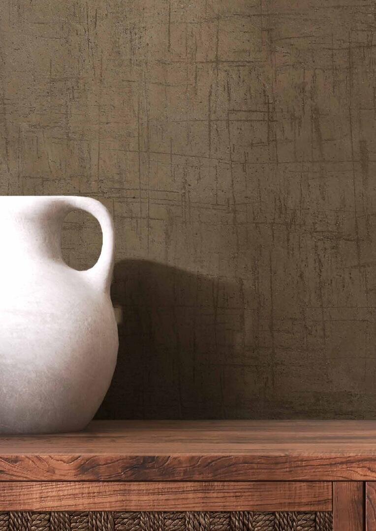A white ceramic vase with a round handle sits on a wooden surface with woven details, against a textured brown wall with a scratched, crisscrossed pattern.