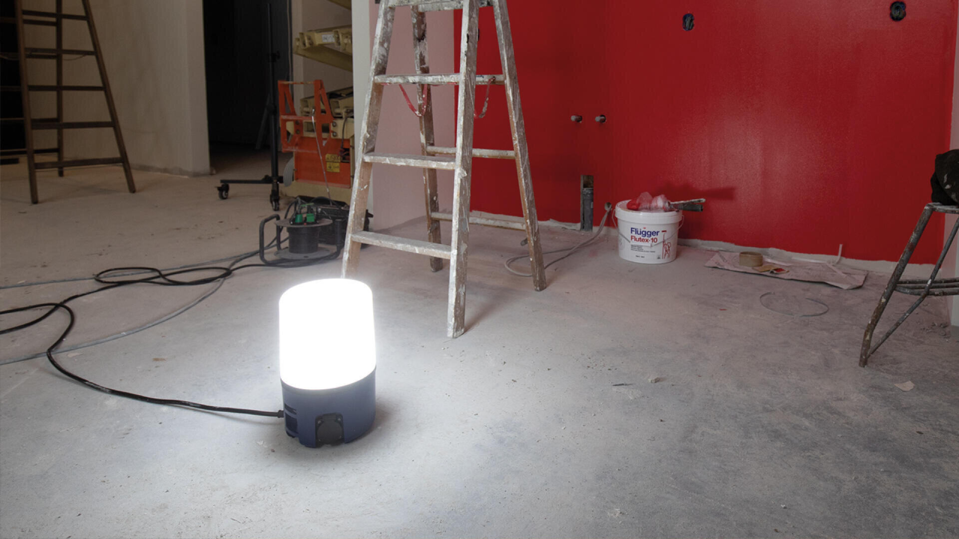 A bright work light sits on a dusty floor in a room under renovation, with ladders, tools, and a red wall visible in the background.