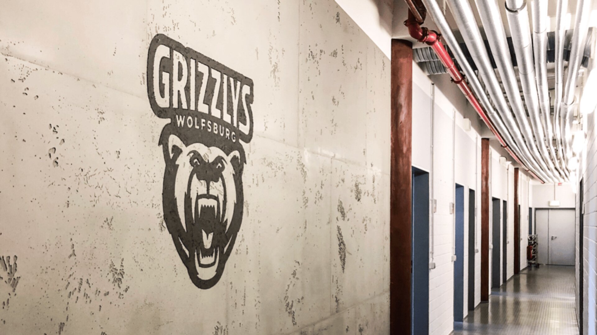 An der Wand eines Betonflurs prangt ein großes Logo mit dem Bären der Grizzlys Wolfsburg, an der Decke verlaufen freiliegende Rohre und auf der gegenüberliegenden Seite befinden sich mehrere Türen.