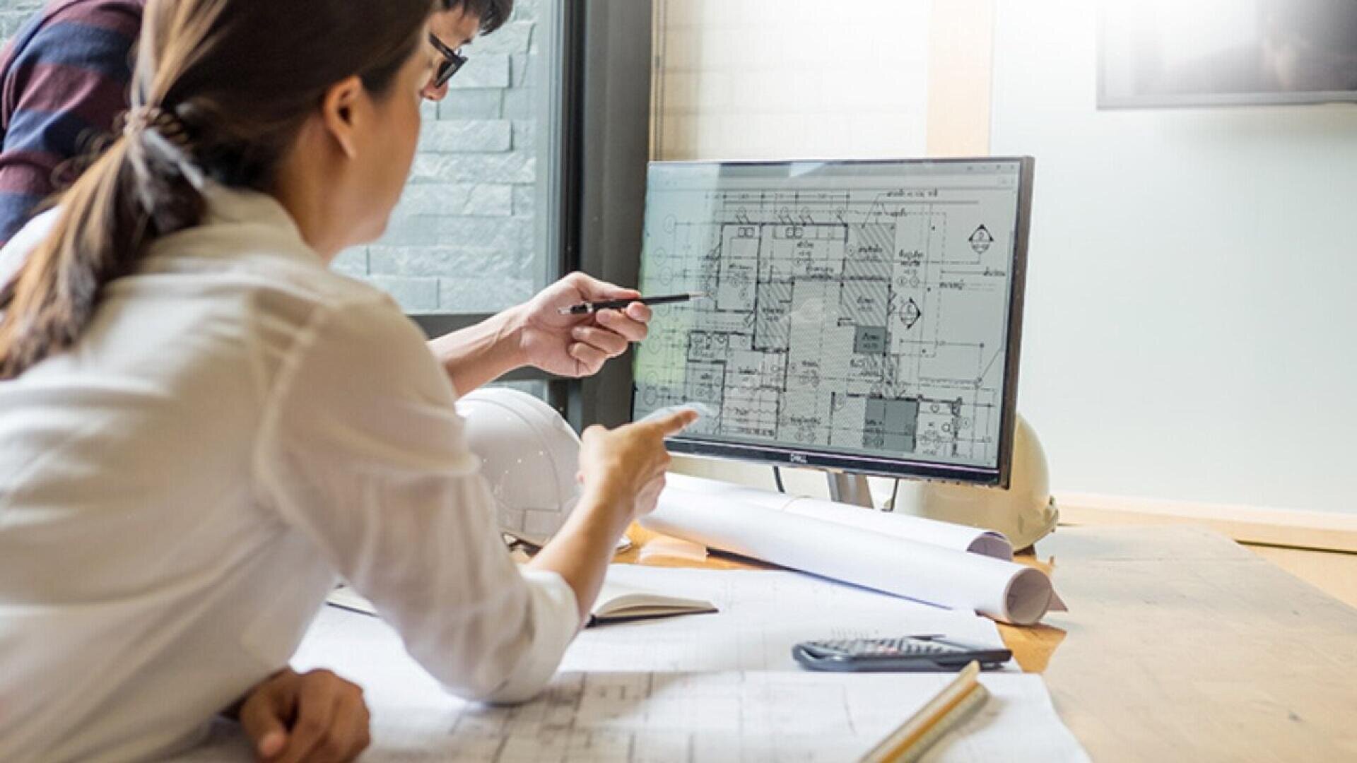 Two people review architectural blueprints displayed on a computer monitor. One holds a pen and points at the screen, while the other gestures. Rolled-up plans, a hard hat, and a calculator are on the desk.