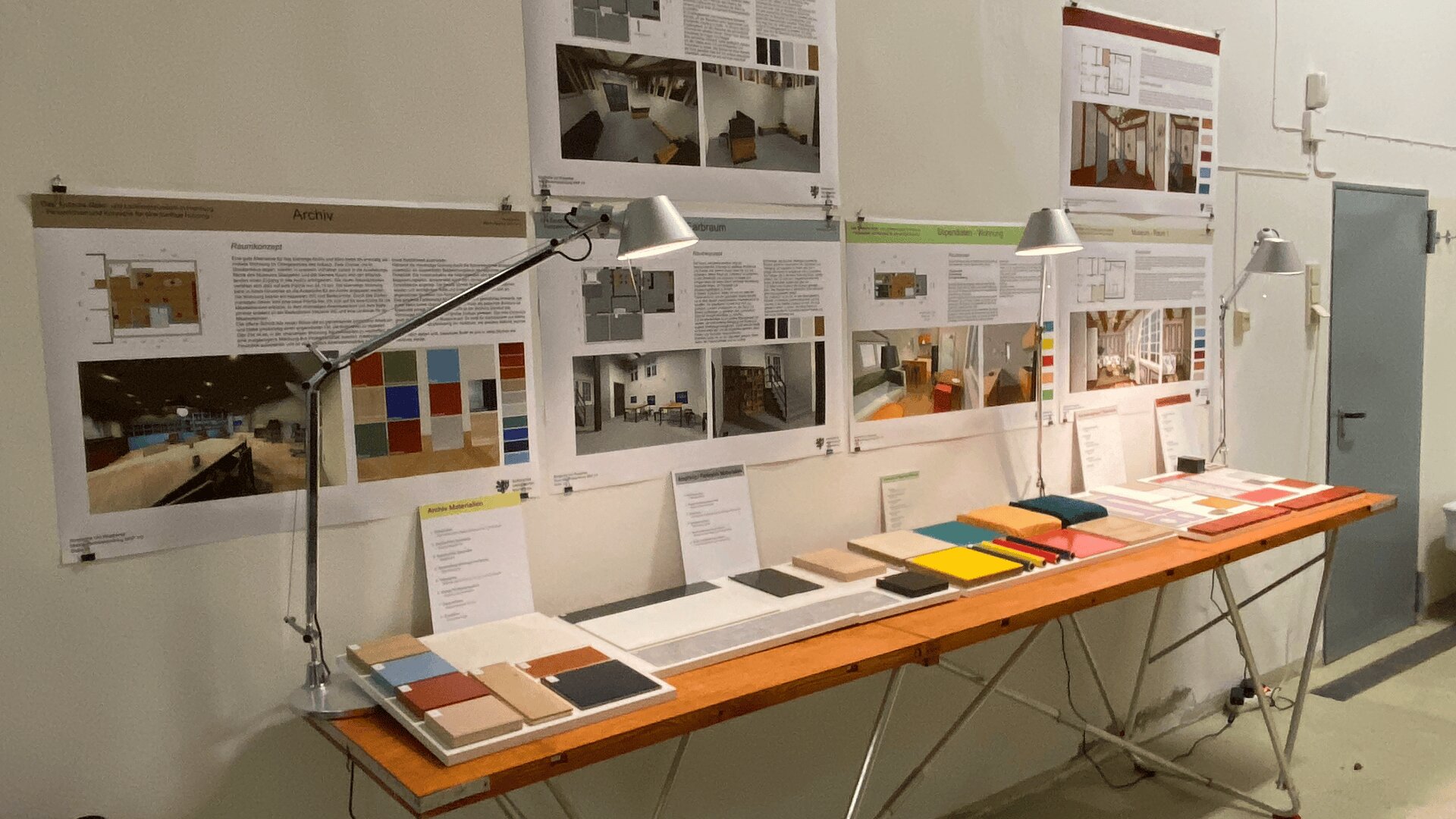 Auf einem Holztisch sind unter Schreibtischlampen Muster farbiger Materialien und Farbmuster ausgestellt, an der weißen Wand dahinter sind Architekturprojekttafeln, Farbpaletten und Designpläne angebracht.