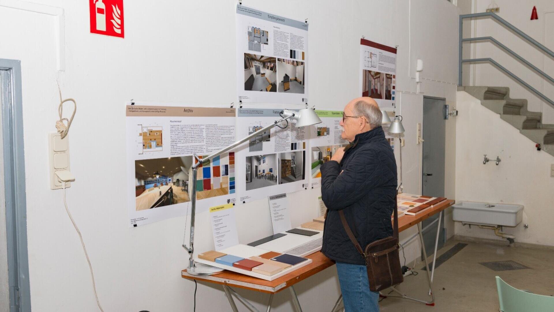 An older man stands in front of a display board with design plans, color samples, and materials, thoughtfully examining the information in a well-lit, industrial-style room.