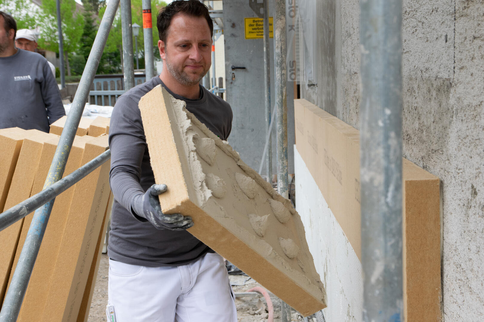 A construction worker holds an insulation panel with adhesive, preparing to attach it to an exterior wall on a building site. Another worker and more insulation panels are visible in the background.