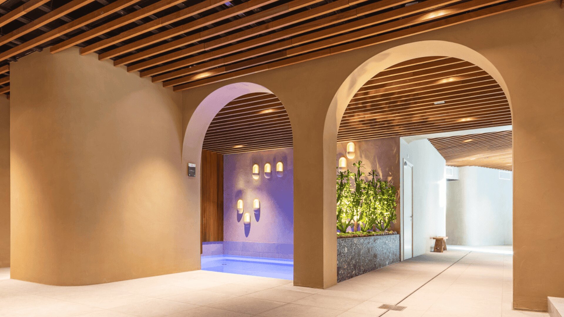 Indoor space with curved archways, wood slat ceiling, and modern decor. A small pool is on the left, illuminated by wall-mounted lights, and a green plant feature lines the hallway on the right. The atmosphere is calm and inviting.