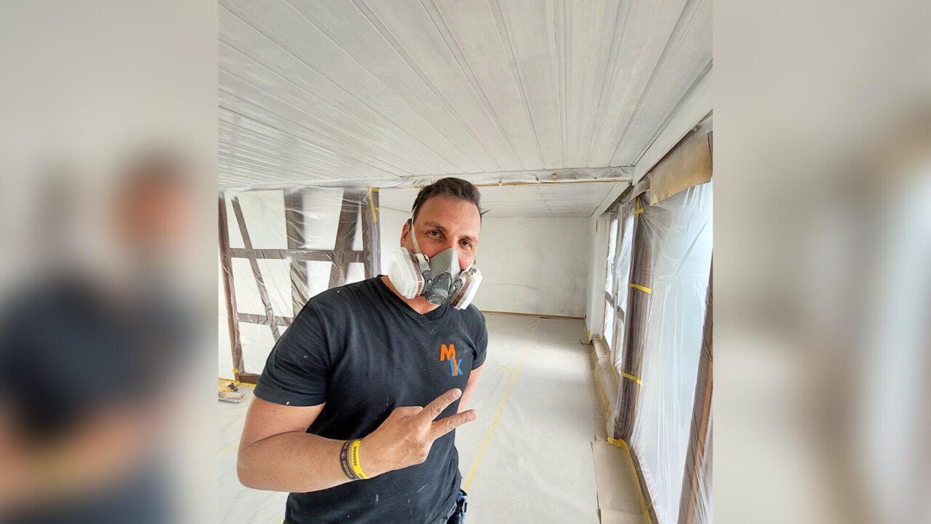 A person wearing a black t-shirt, wristbands, and a protective mask flashes a peace sign while standing in a room under renovation with plastic sheets covering the walls and floor.