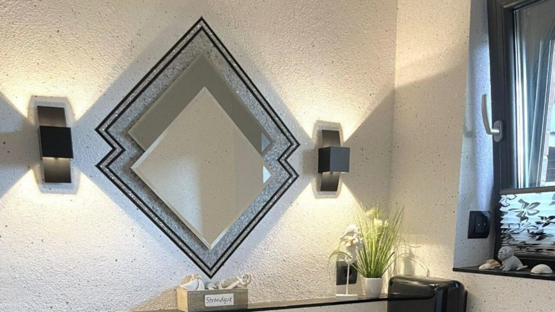 A geometric diamond-shaped mirror is mounted on a textured white wall between two black modern wall lights. Below, a small table holds a potted plant and tissue box. A window with frosted glass is on the right.