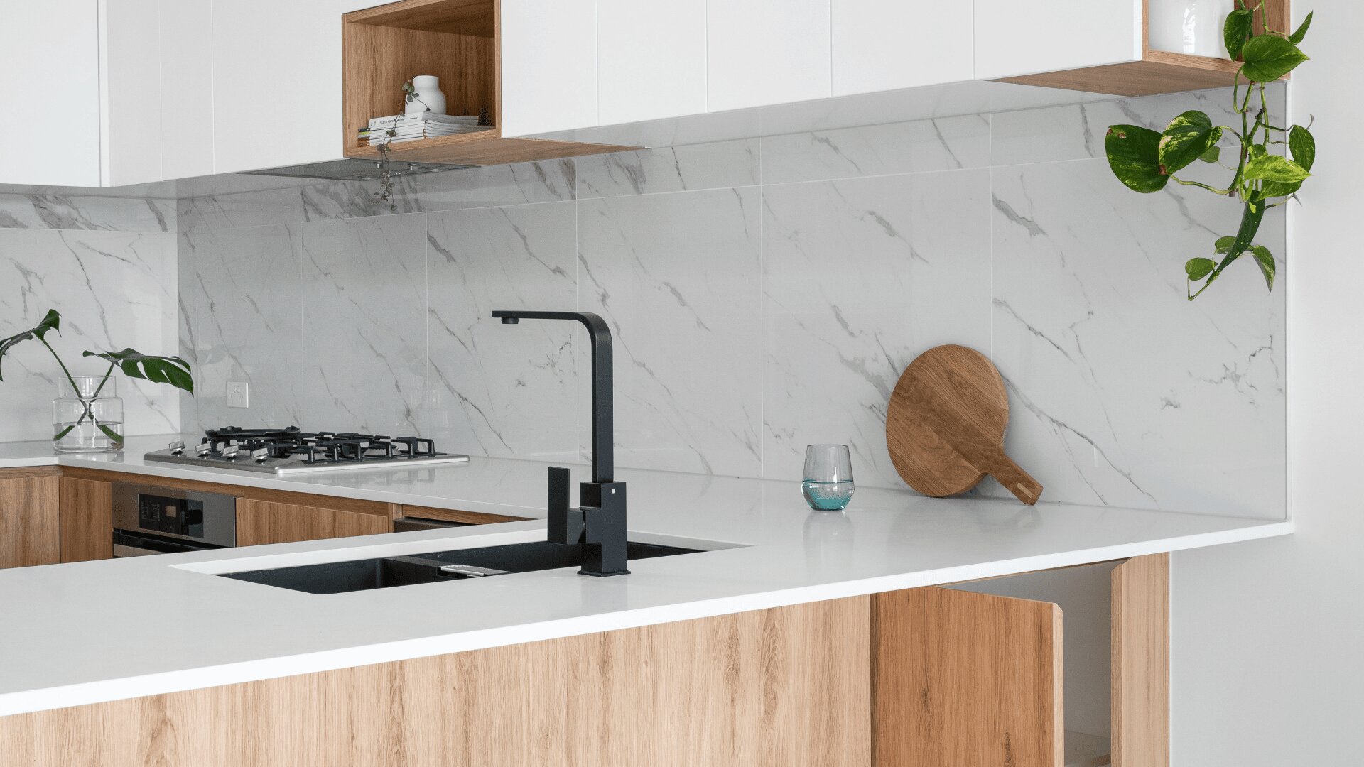 Moderne Küche mit weißer Marmorrückwand, Holzschränken, schwarzem Wasserhahn, eingebautem Gasherd, Topfpflanzen, einem runden Holzschneidebrett und einem Glas auf der Arbeitsplatte.
