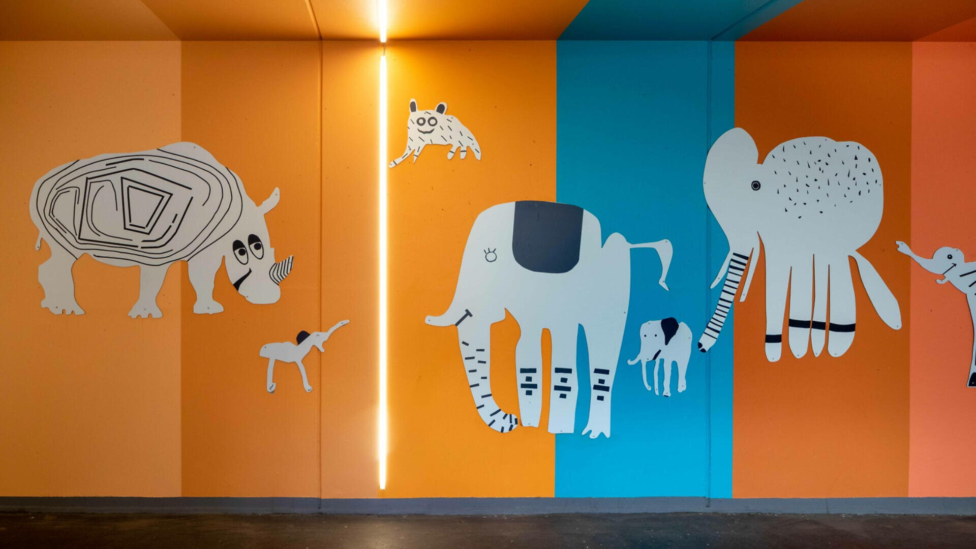 Ein Wandgemälde auf einer farbenfrohen orange-blauen Wand zeigt verspielte, cartoonartige Tiere, darunter ein Nashorn, Elefanten, eine Hyäne und einen Affen, alle mit einfachen schwarzen und weißen Linien und Formen gezeichnet.