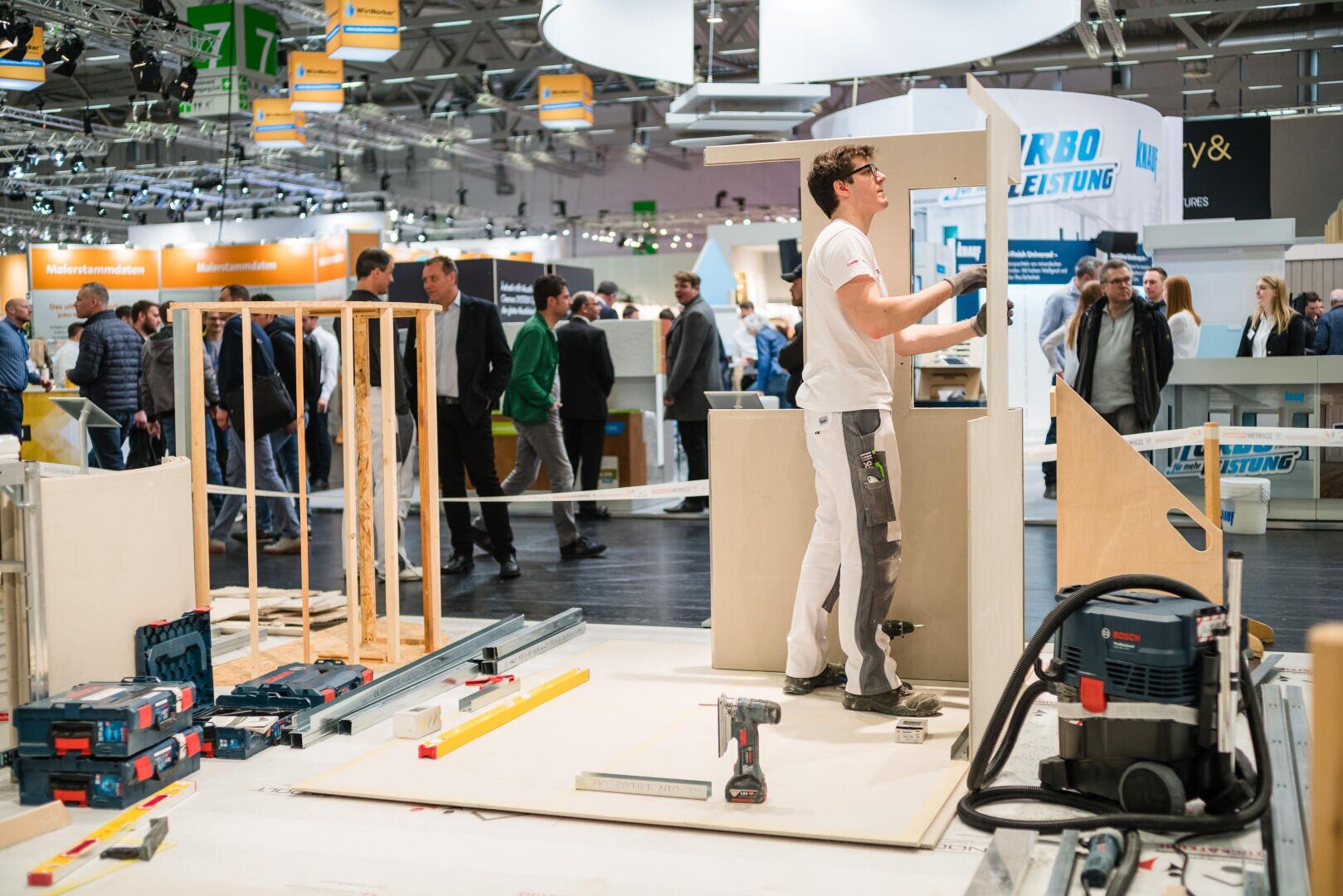Ein Mann in Arbeitskleidung demonstriert auf einer Messe seine Tischlerkunst und montiert Holzplatten. Werkzeuge und Geräte liegen verstreut um ihn herum, während Menschen in der geschäftigen Messehalle umhergehen und zuschauen.