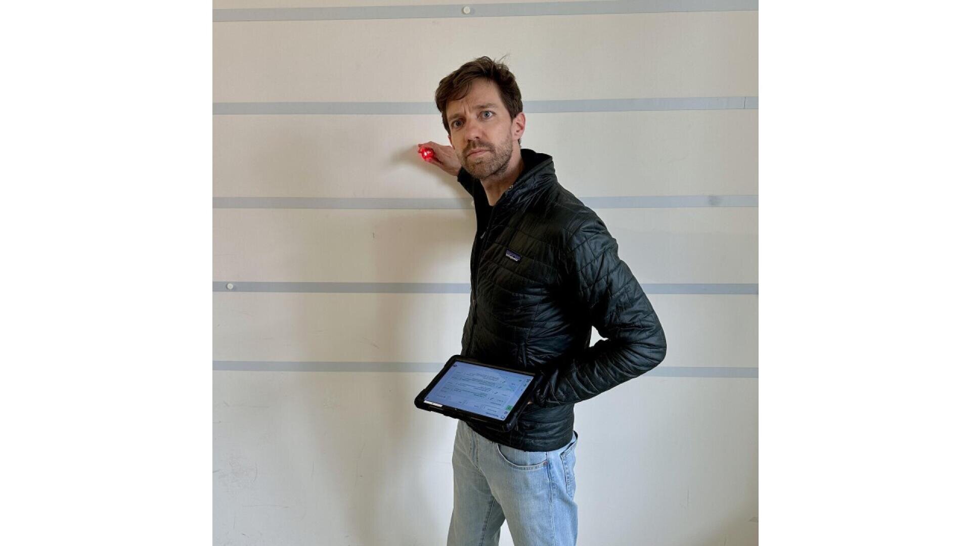 A man in a black jacket holds a tablet in one hand and touches a white wall with horizontal gray lines with the other, looking at the camera.