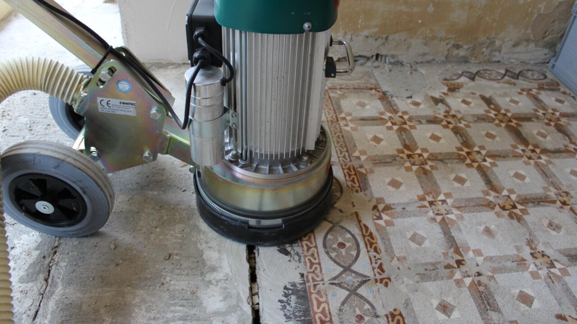 A floor grinder machine is being used to remove old, patterned tiles from a floor, exposing the bare concrete beneath. The process is partially complete, showing both the original tiles and stripped area.