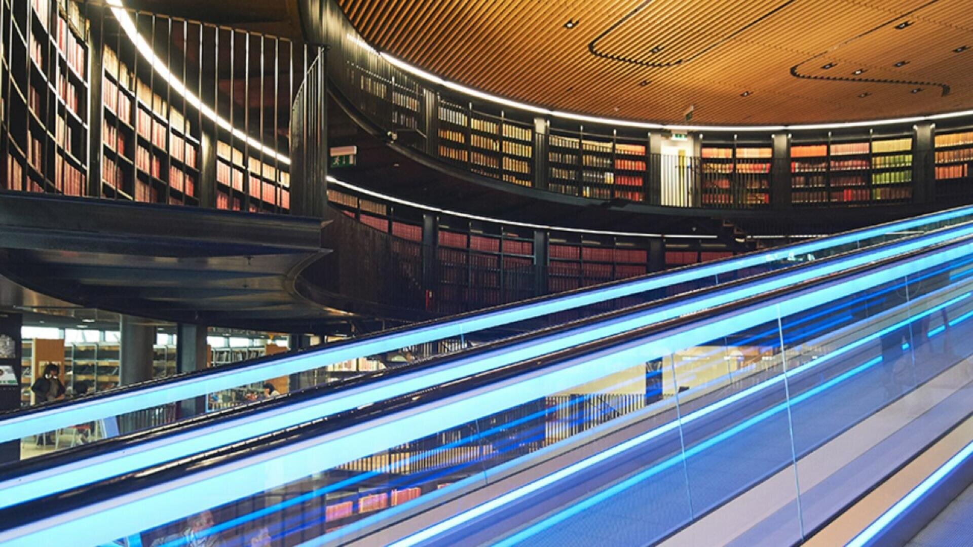 Curved bookshelves filled with colorful books line the walls of a modern library. A glass escalator with blue lighting runs diagonally through the foreground, creating a dynamic, futuristic atmosphere.