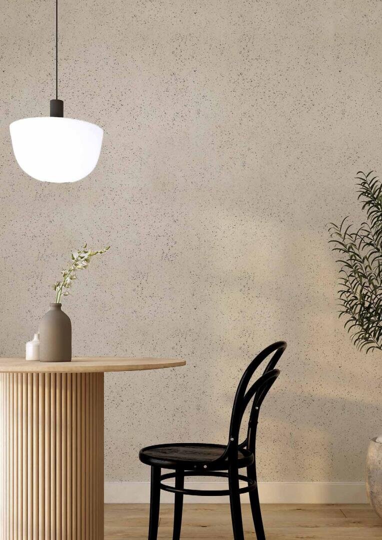 A modern minimalist dining area featuring a round wooden table with a vase holding a single branch, a black chair, a white pendant light, and a potted plant against a speckled beige wall.