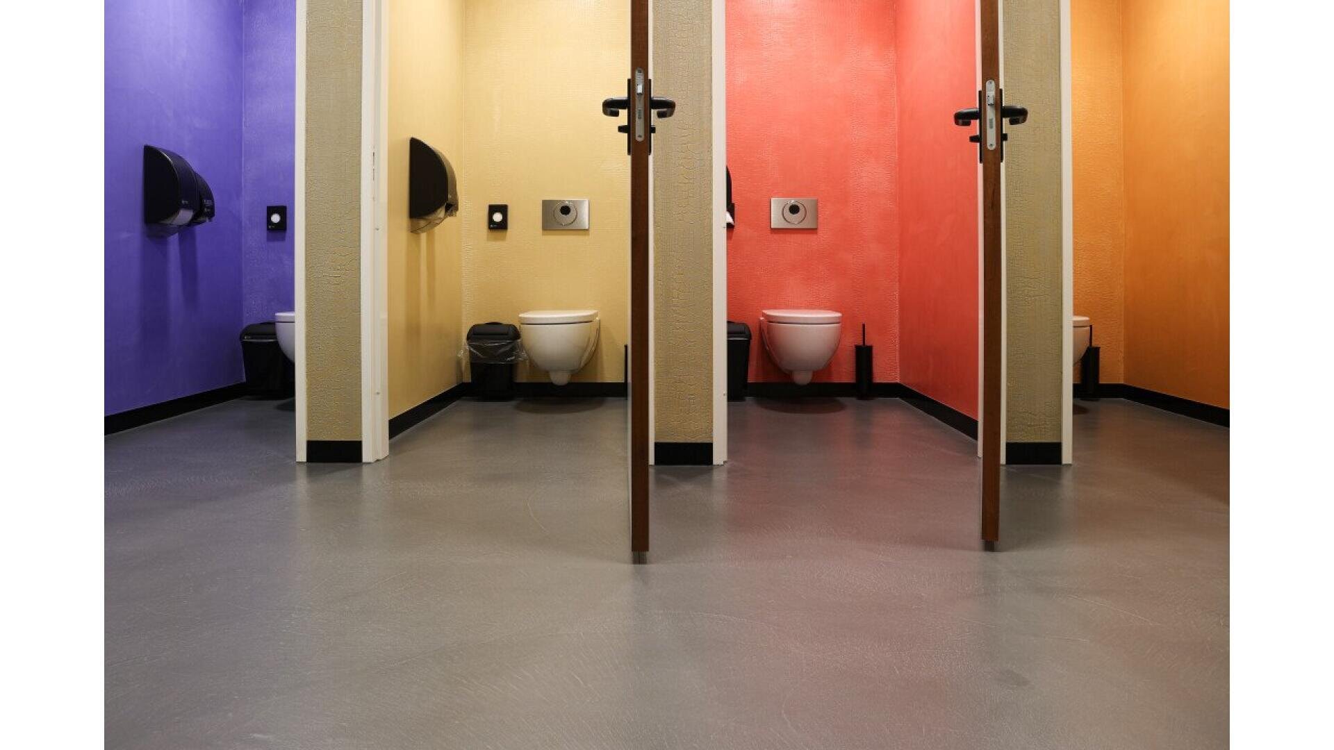 Three bathroom stalls with doors open, each painted a different color: purple, yellow, and red. Each stall has a toilet, a wall-mounted toilet paper dispenser, and a waste bin on the floor.