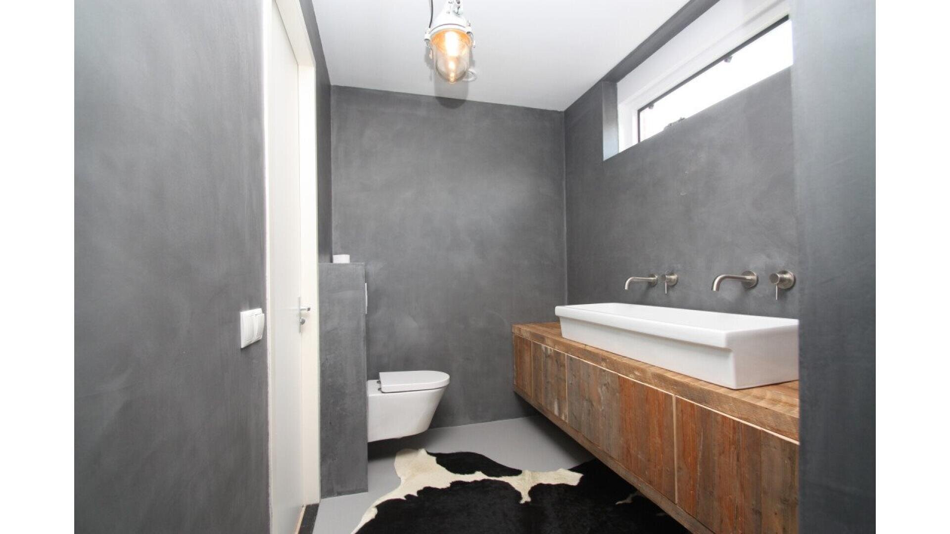 Minimalist bathroom with gray walls, a floating wooden vanity with a long white rectangular sink, two wall-mounted faucets, a wall-hung toilet, a window above the sink, and a black-and-white cowhide rug on the floor.