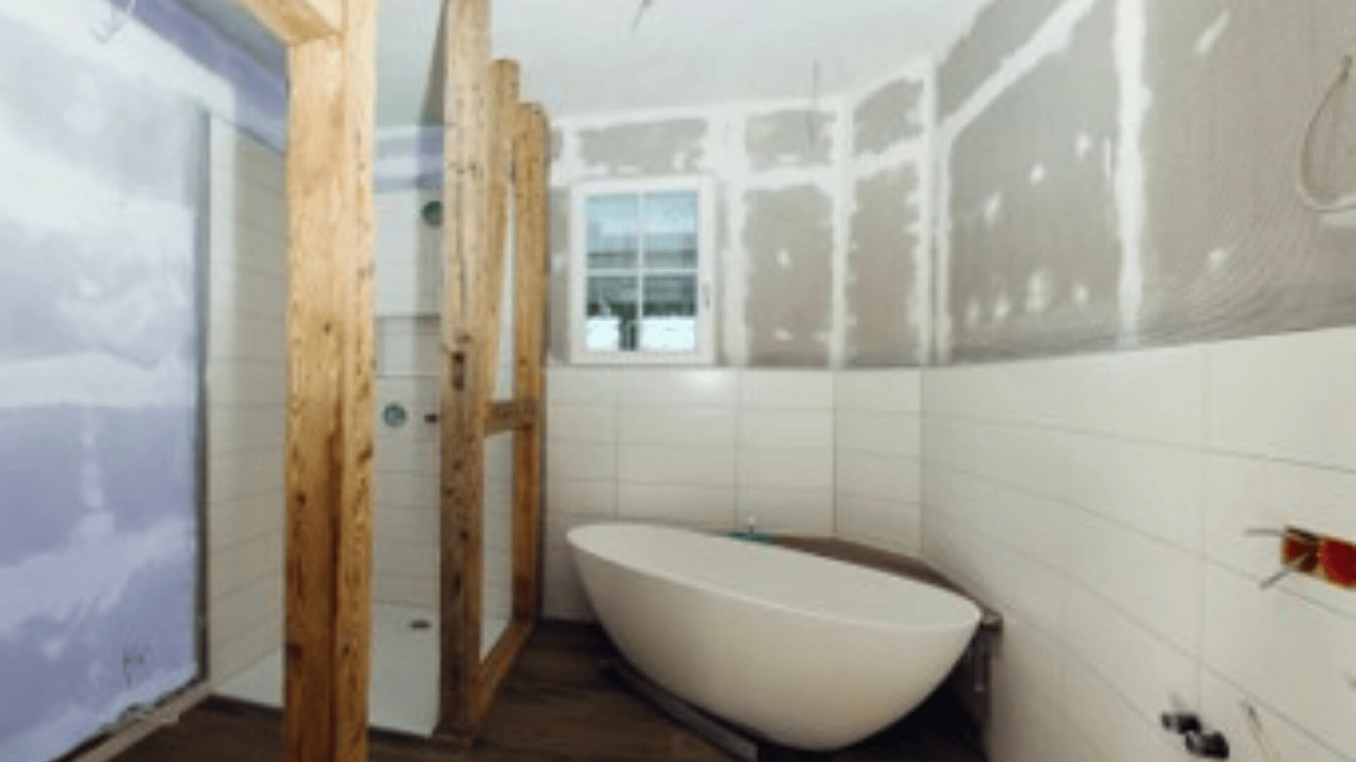 Ein Badezimmer im Umbau mit unfertigen Wänden, einer freistehenden weißen Badewanne, gefliesten unteren Wänden, freiliegenden Holzbalken und einem kleinen Fenster.