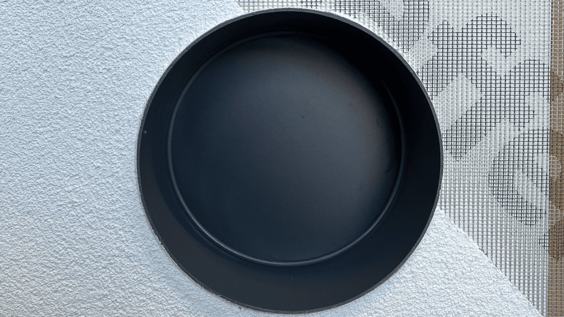A round, empty black pan sits on a textured white surface with a mesh screen and patterned shadow in the background.