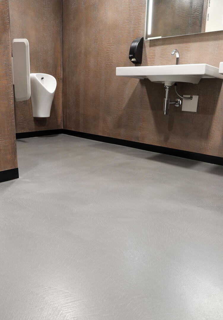 Eine saubere, moderne öffentliche Toilette mit strukturierten braunen Wänden, einem weißen Urinal, einem weißen Wandwaschbecken mit freiliegenden Rohren, einem Seifenspender und einem grauen Boden.