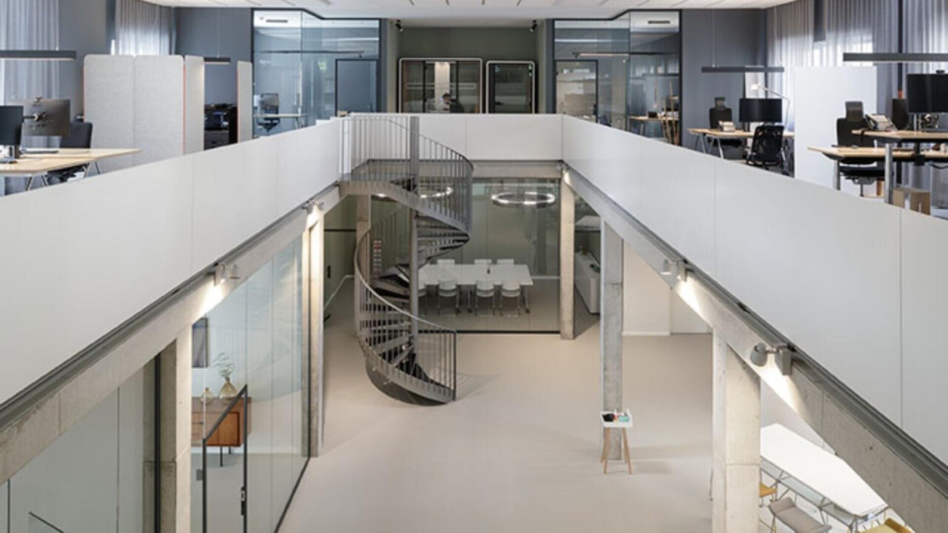 Modern open-plan office with glass-walled meeting rooms on the ground floor, workstations on the upper level, and a central spiral staircase. The space is bright with minimalist decor and neutral colors.