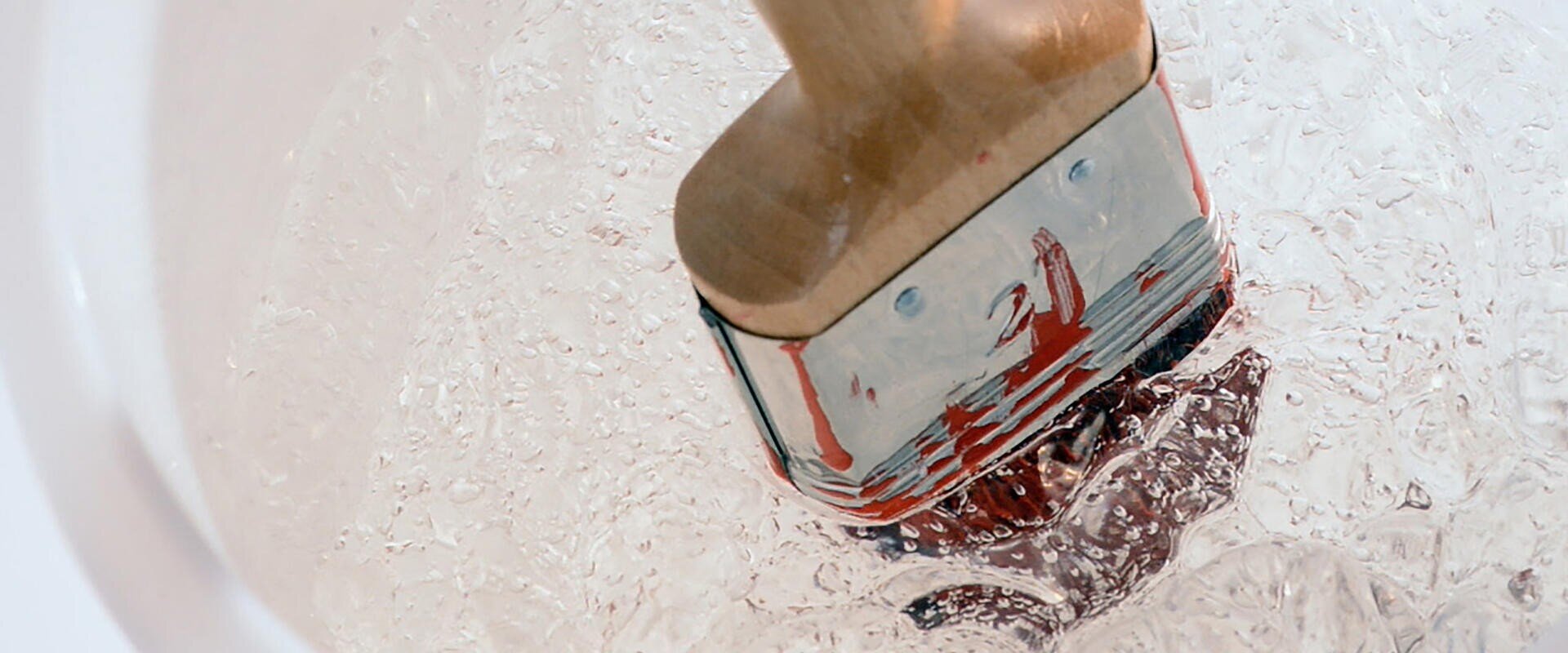 A paintbrush with red paint on its bristles is being rinsed in a clear container of water, creating swirling patterns and bubbles.