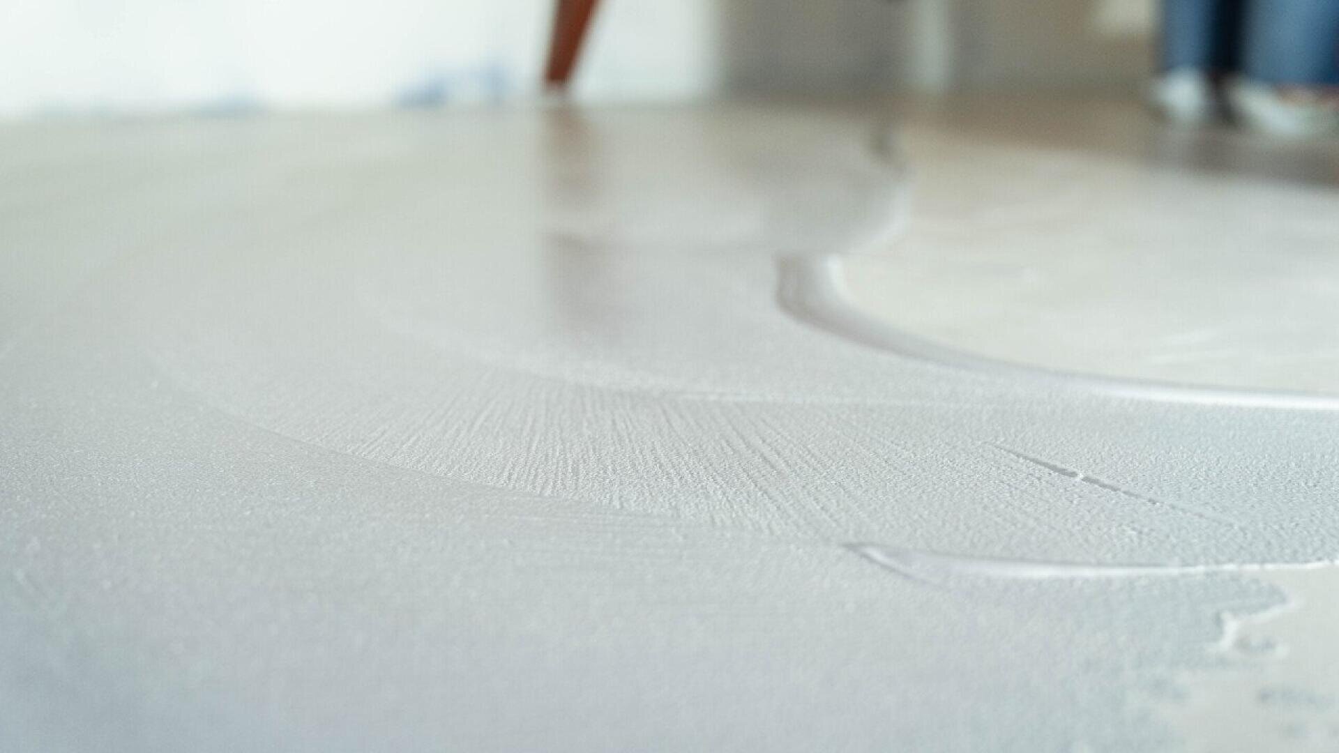 Close-up of a smooth, wet layer of white paint or plaster being applied to a surface, with faint brush or trowel marks visible. The background is slightly blurred.