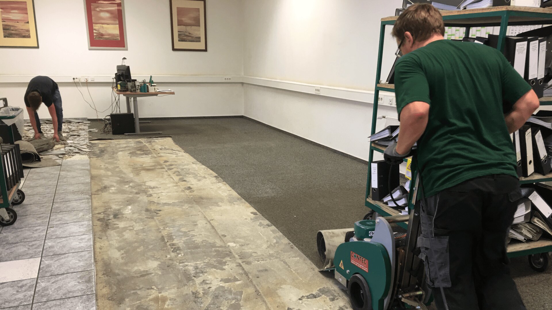 Zwei Arbeiter entfernen alten Bodenbelag in einem Büro. Eine Person hebt Fliesen an der Wand an, während eine andere eine Bodenabziehmaschine bedient. Im Hintergrund sind ein Regal mit Ordnern und Bürogeräten zu sehen.
