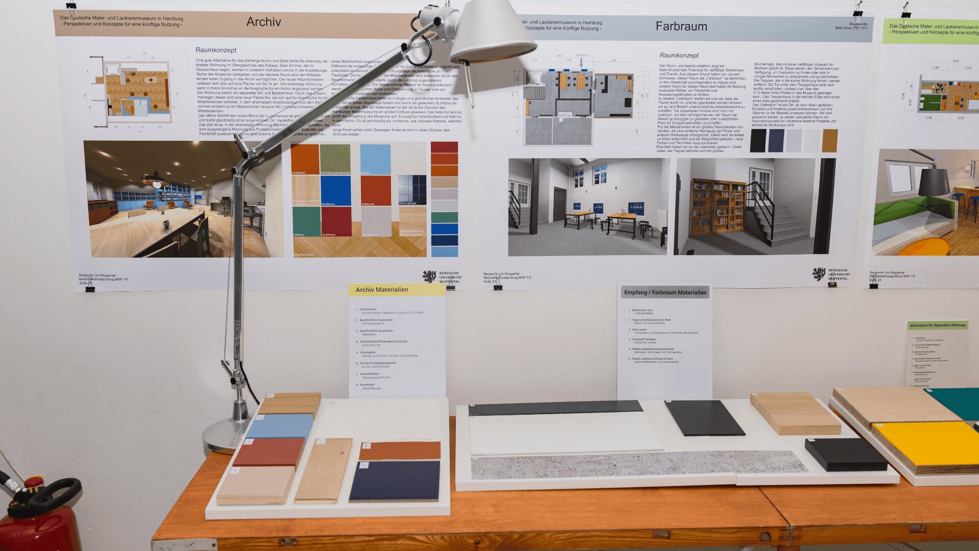 Ein Arbeitsplatz mit Schreibtischlampe, Materialmustern und Infotafeln an der Wand, die Raumkonzepte, Farbpaletten und Einrichtungsideen zeigen. Links auf dem Boden steht ein Feuerlöscher.