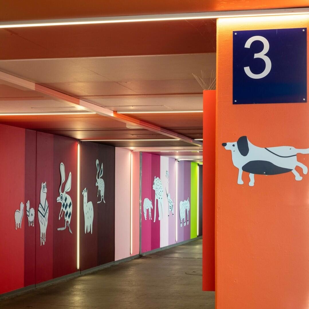 Auf der mit orangefarbenen Wänden gekennzeichneten Ebene 3 des Parkhauses ist eine weiß-schwarze Hundeillustration zu sehen; an den bunten Wänden sind verschiedene weiße Tierzeichnungen zu sehen, darunter eine Biene, eine Katze und ein Schaf.