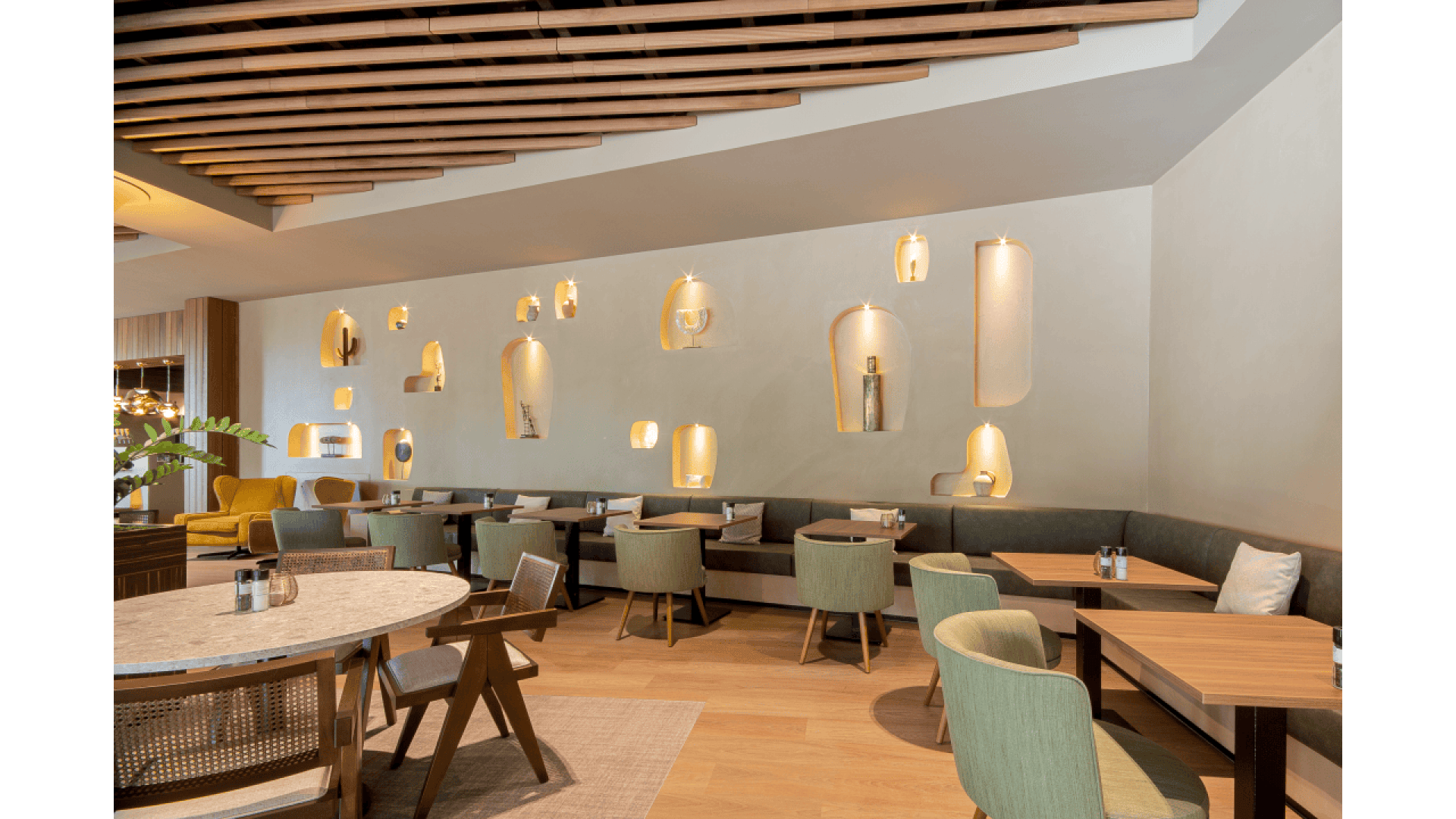 Modern restaurant interior with wooden tables, cushioned chairs, and a curved booth along the wall. The white wall features decorative alcoves with lights and art pieces. The ceiling has wooden slats. The space looks bright and stylish.