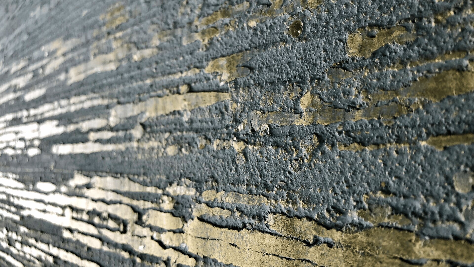 Nahaufnahme einer strukturierten Wand mit unregelmäßigen horizontalen Rillen und Streifen, die eine Kombination aus rauen grauen und goldfarbenen Oberflächen aufweist. Licht hebt die Tiefe und die metallischen Akzente der Wandoberfläche hervor.