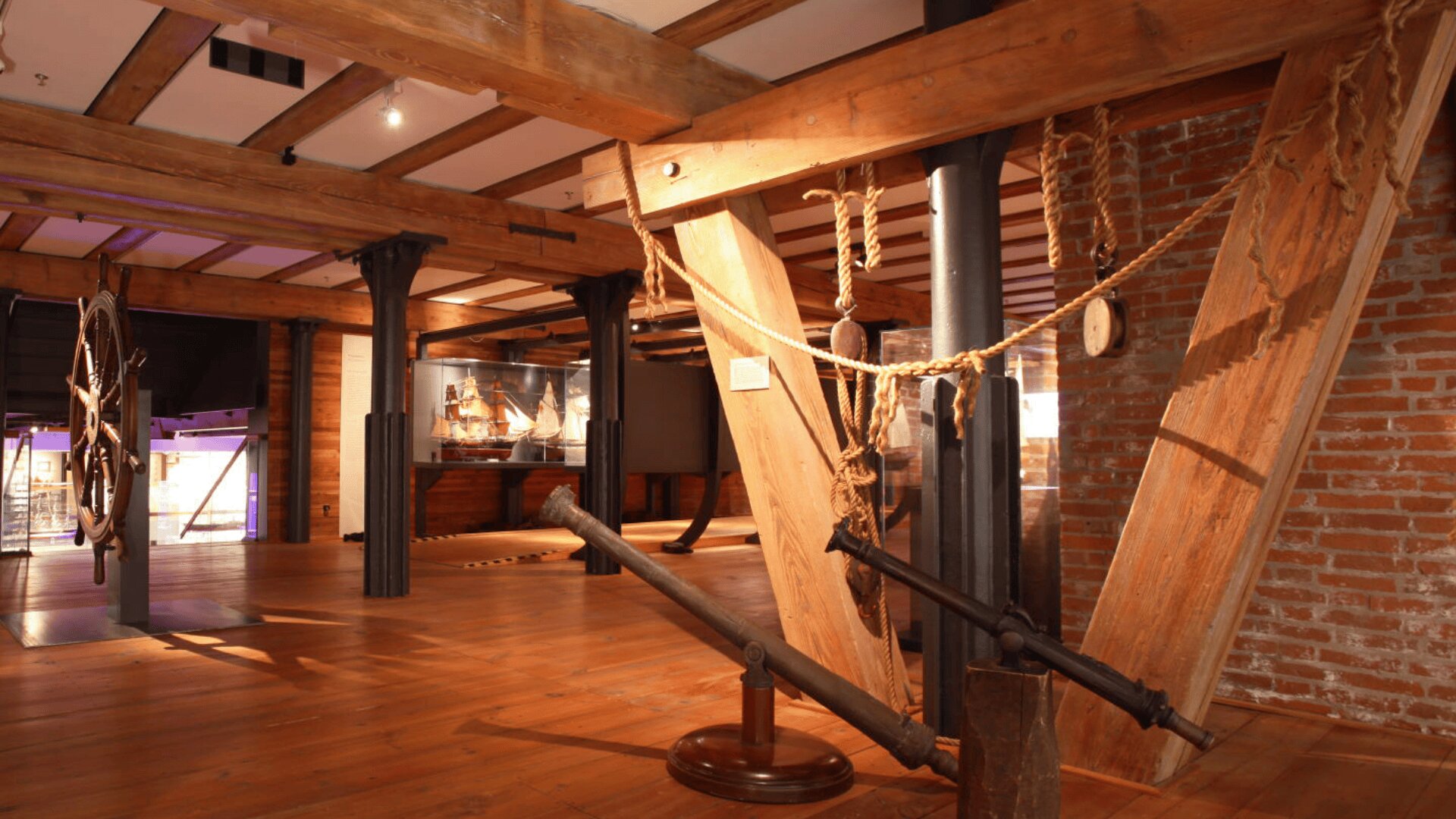 Die hölzerne Innenausstattung eines Schifffahrtsmuseums mit großen Holzbalken, einer Ziegelwand, nautischen Seilen und einem Schiffsanker. Im Hintergrund ist eine Vitrine mit Schiffsmodellen zu sehen.