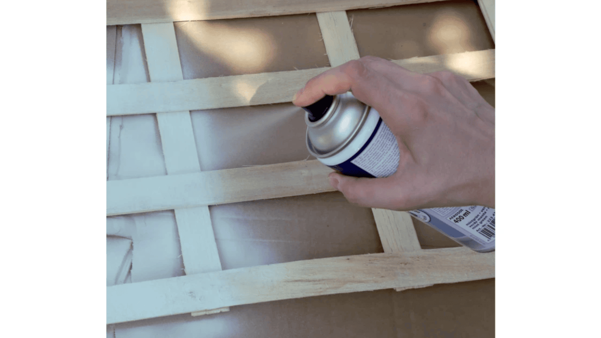 A hand is holding a can of spray paint, applying white paint to a wooden lattice placed on top of cardboard.