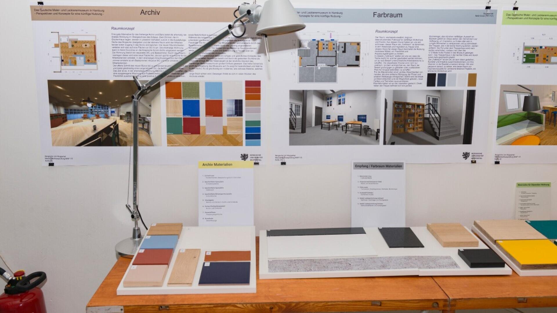 A display table with various material and color samples under a desk lamp. Behind the table are posters showing room designs, color concepts, and material information, attached to a white wall.