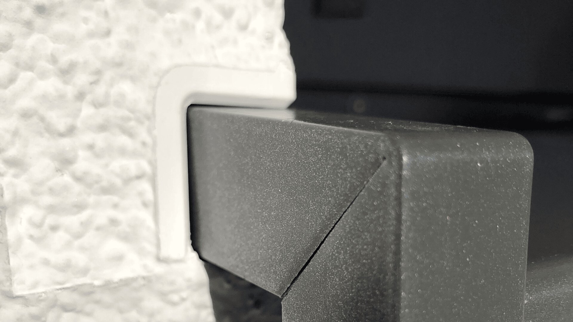 Nahaufnahme einer strukturierten weißen Wandecke mit einem darum angebrachten Kantenschutz aus grauem Kunststoff oder Metall, der einen scharfen Kontrast zwischen den Oberflächen erzeugt und die geometrischen Formen hervorhebt.