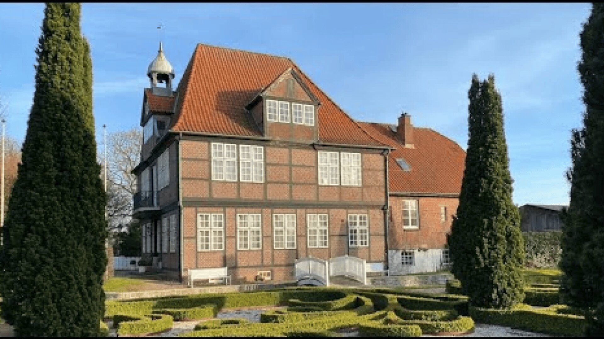 A traditional half-timbered house with a red-tiled roof and many windows, surrounded by neatly trimmed topiary trees and a manicured garden under a blue sky.