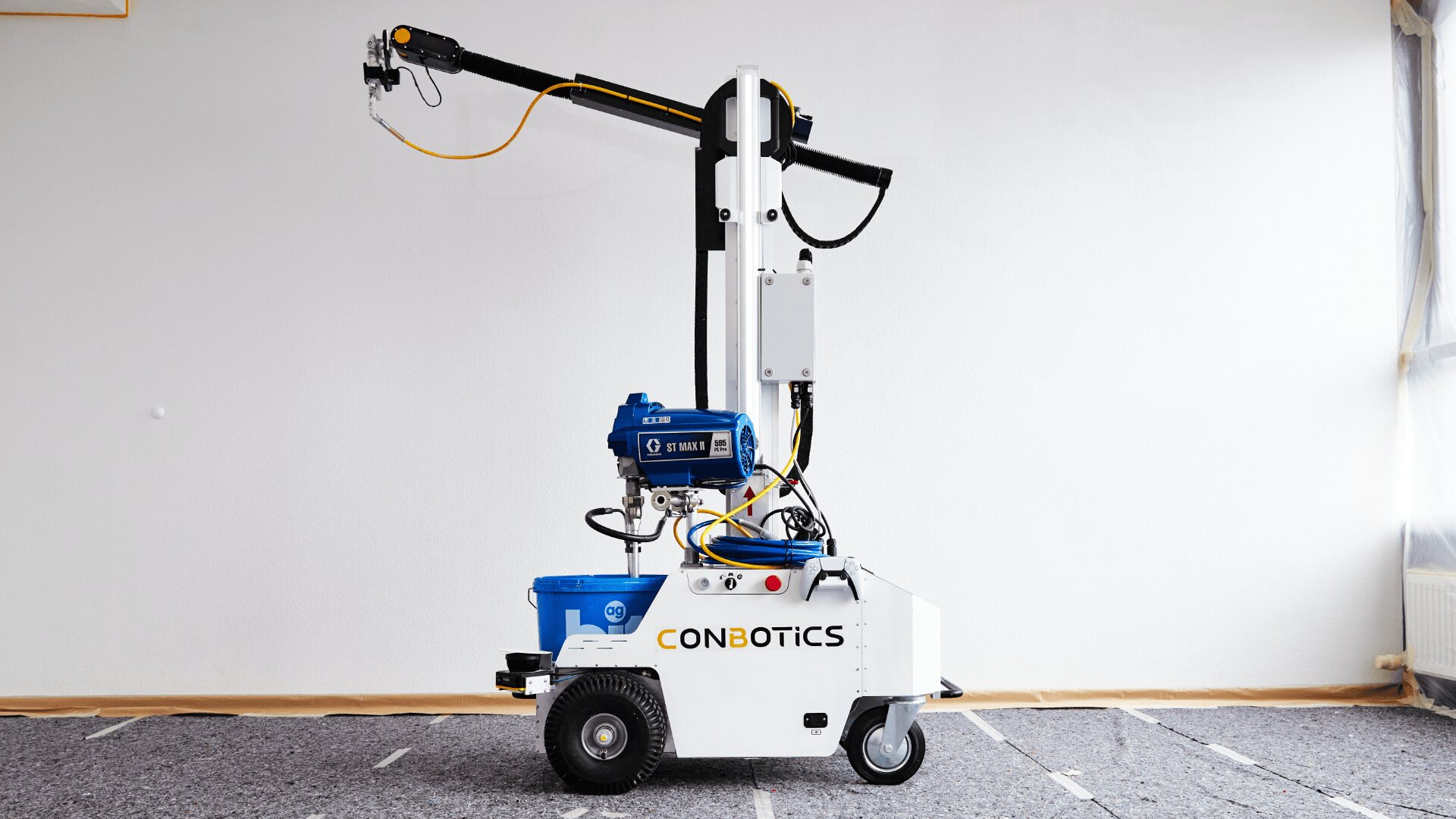 Ein weißer, kastenförmiger Roboter mit der Aufschrift „CONBOTICS“ mit Rädern, einem blauen Motor und einem langen, aufrechten Arm steht auf einem mit Teppich ausgelegten Boden vor einer schlichten weißen Wand.