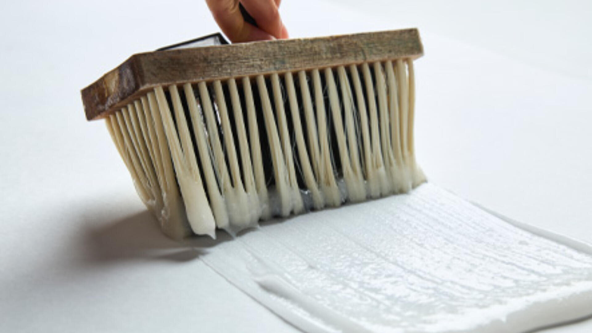 Eine Hand hält einen großen Pinsel und verteilt damit eine Schicht weißen Klebers oder Klebstoffs auf einer flachen Oberfläche.