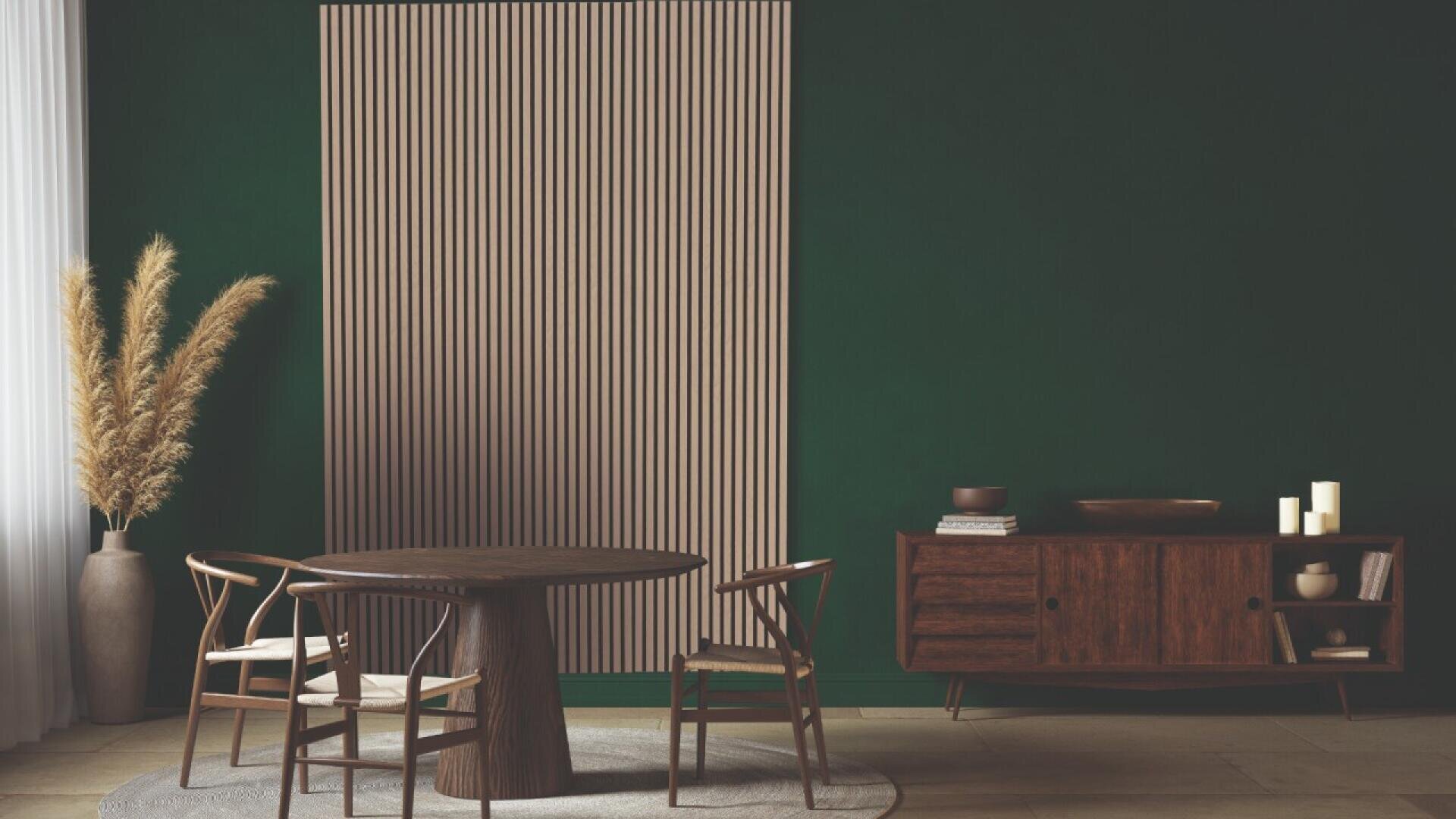 A modern dining room with dark green walls, a round wooden table, four wooden chairs, a sideboard with decor, tall pampas grass in a vase, and a patterned wall panel behind the table.