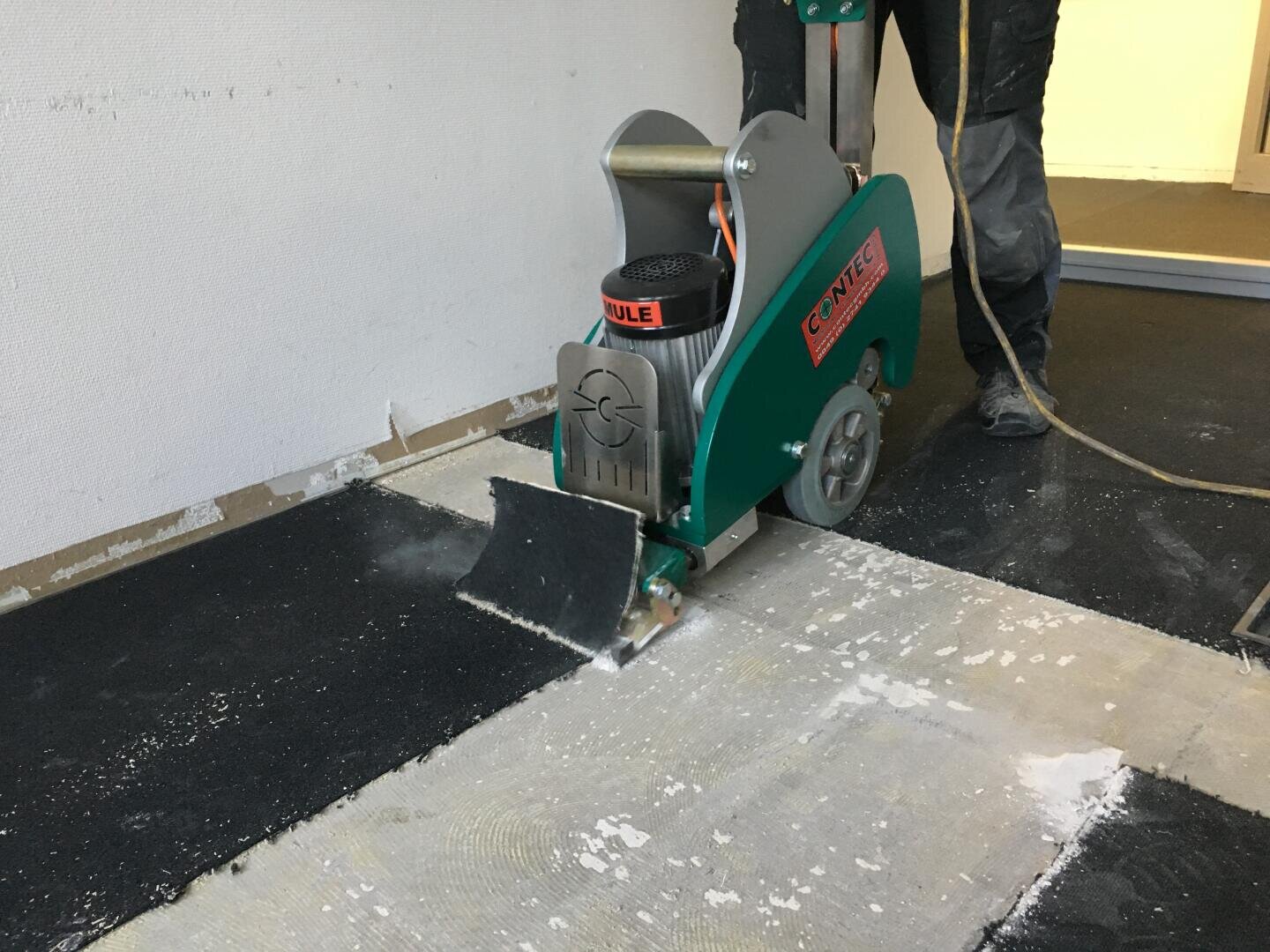 Eine Person bedient eine umweltfreundliche Bodenabziehmaschine, um alte Bodenbeläge aus einem Raum zu entfernen, wobei die darunterliegende Oberfläche freigelegt und Schmutz zurückgelassen wird.